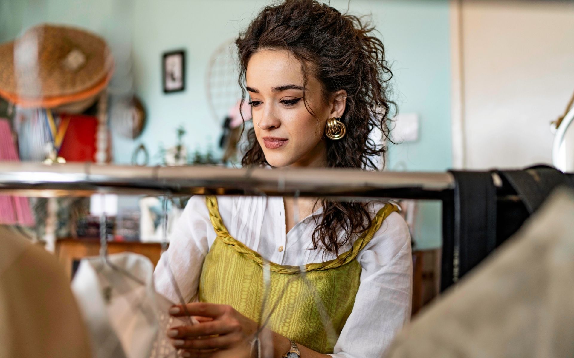 Smiling woman browsing through clothes on a clothing rack in a charity shop, wearing a white shirt and a yellow corset top, enjoying a sustainable shopping experience.