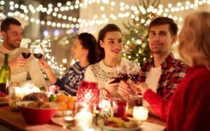 Five friends celebrating Christmas dinner, wearing festive Christmas jumpers, clinking red wine glasses in a toast, with smiles and laughter around the table