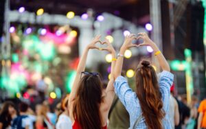 Two friends at a concert with a blurry background, holding their hands up in the shape of hearts, surrounded by colourful lights and a festive atmosphere