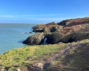 Scenic hike near Dublin: Rugged coastline with a scenic trail winding along the cliffs. A hidden cave nestled in the rocky shore. Clear blue skies overhead, reflecting in the sparkling blue waters below.