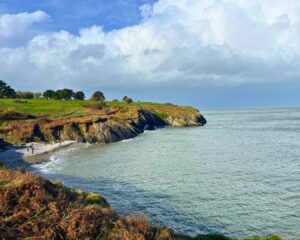 Scenic hike near Dublin: Dramatic coastline where rugged cliffs covered in greenery meet the deep blue sea. Below, a small sandy beach nestles between the cliffs, with a family playing near the water. The sky is bright blue with scattered white clouds.