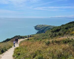 Scenic hike near Dublin: Stunning view of Howth Cliff Walk, with a glimpse of Howth Lighthouse in the distance. People are walking along the trail on a sunny day, with clear blue skies overhead.