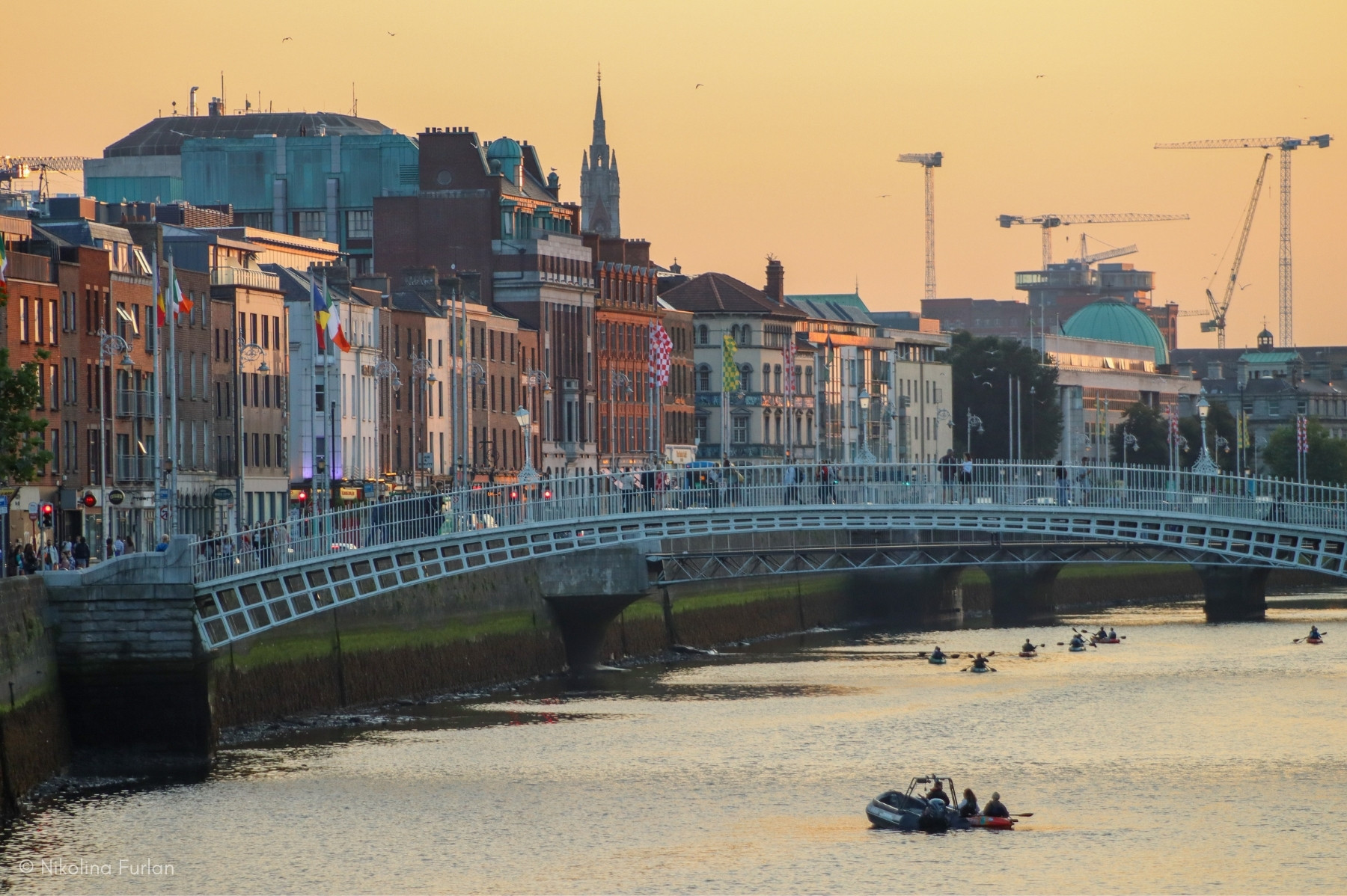 Ha’penny Bridge over the River Liffey in Dublin at sunset, with people walking across and boats paddling below, set against a cityscape with cranes and historic buildings.