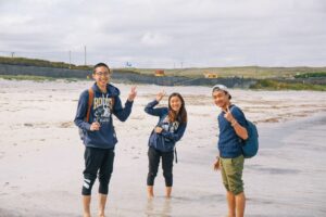 Three Atlas Language School students smiling and posing with peace signs while standing barefoot in the shallow waters of a beach on the Aran Islands