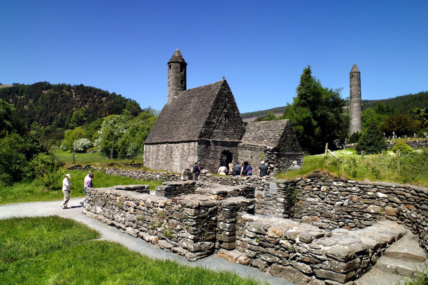 Visitors exploring the historic stone church and round tower at Glendalough Monastic Site, surrounded by green hills and forest under a clear blue sky in County Wicklow, Ireland.