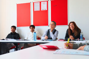 Adult students participating in an English language class at Atlas Language School Liverpool, seated in a bright classroom with red bulletin boards and study materials.